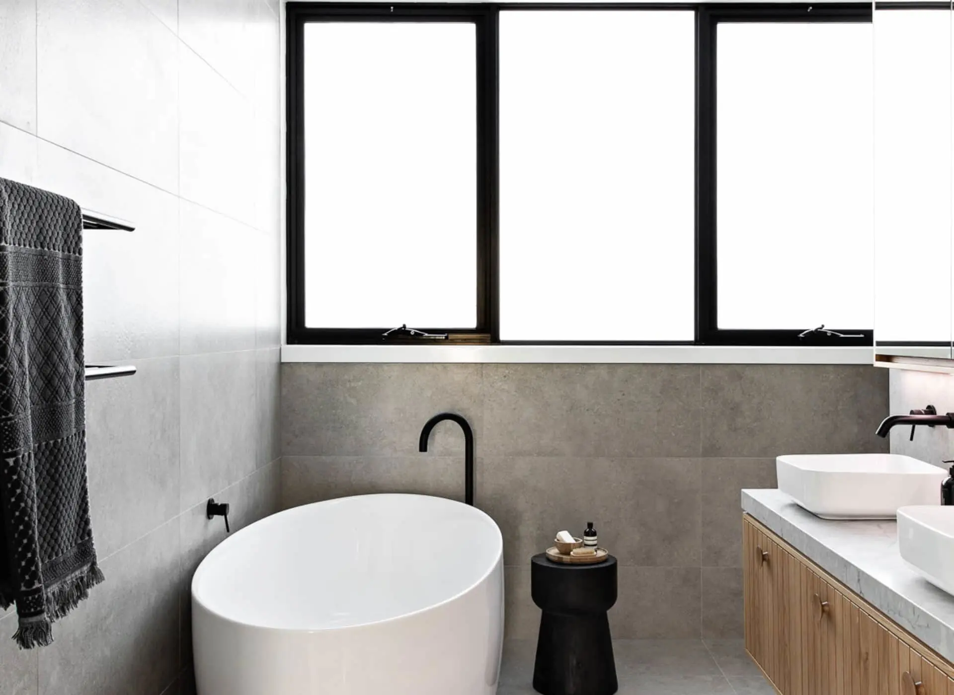 Modern bathroom with white freestanding tub, black fixtures, gray tile walls, wooden vanity, and large windows.
