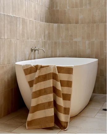 Modern white freestanding bathtub with brown striped towel in a beige tiled bathroom with wall-mounted faucet.