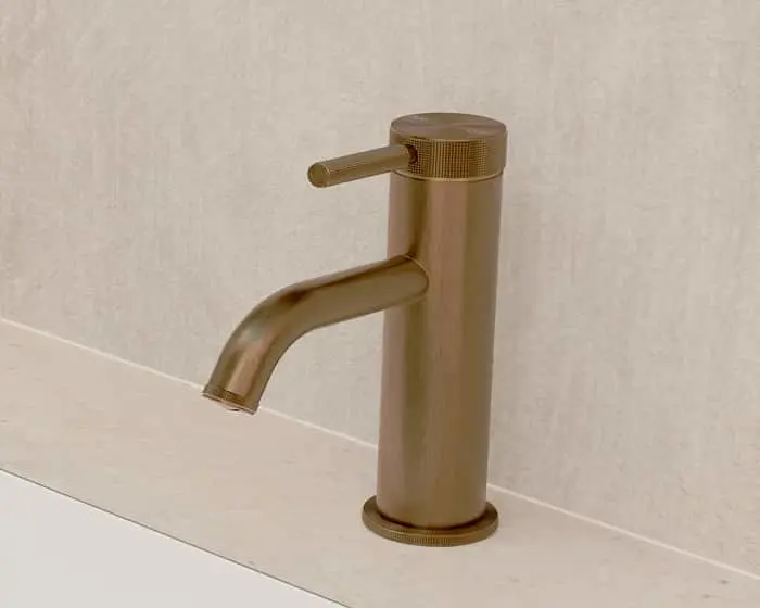 Modern bronze bathroom faucet with cylindrical body and curved spout mounted on a white countertop against beige wall.