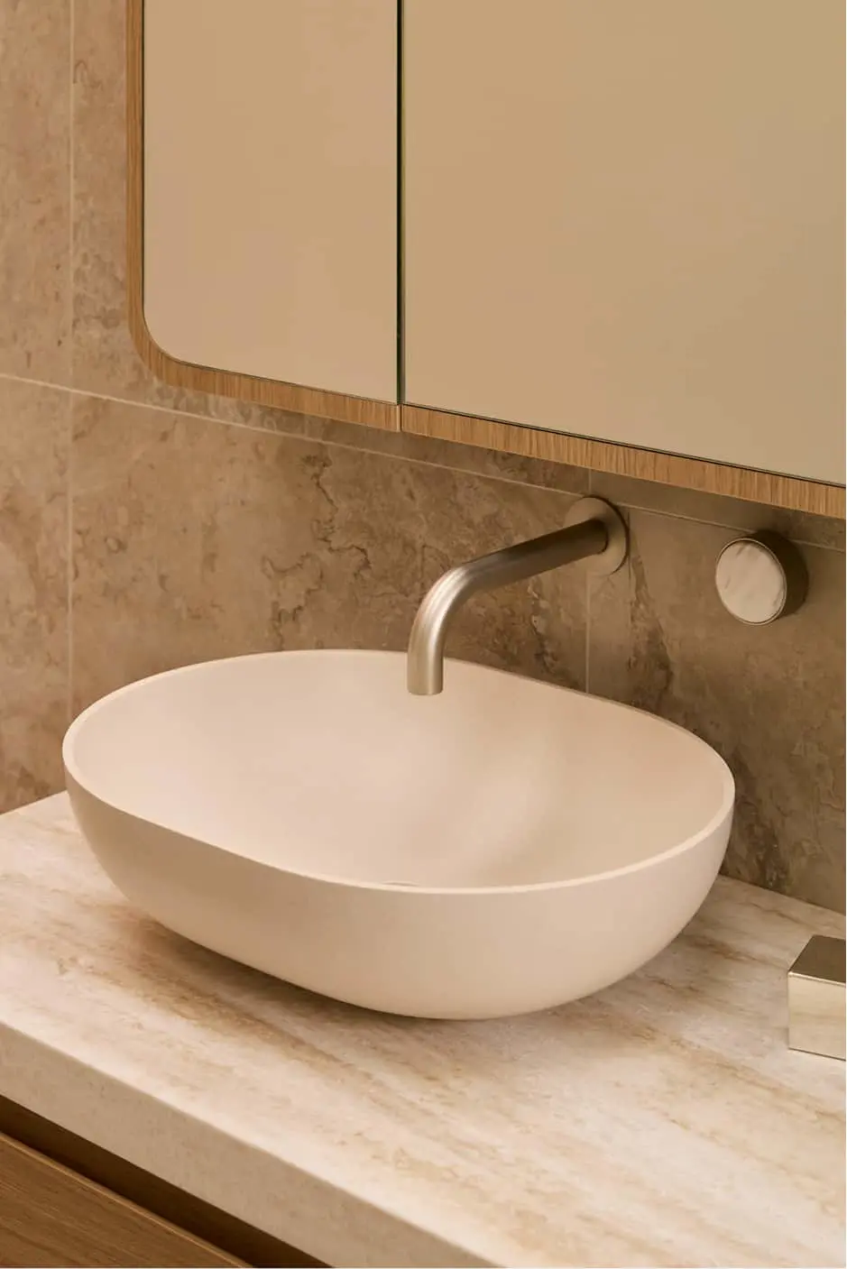 White oval vessel sink on stone countertop with wall-mounted brushed metal faucet and mirror above.