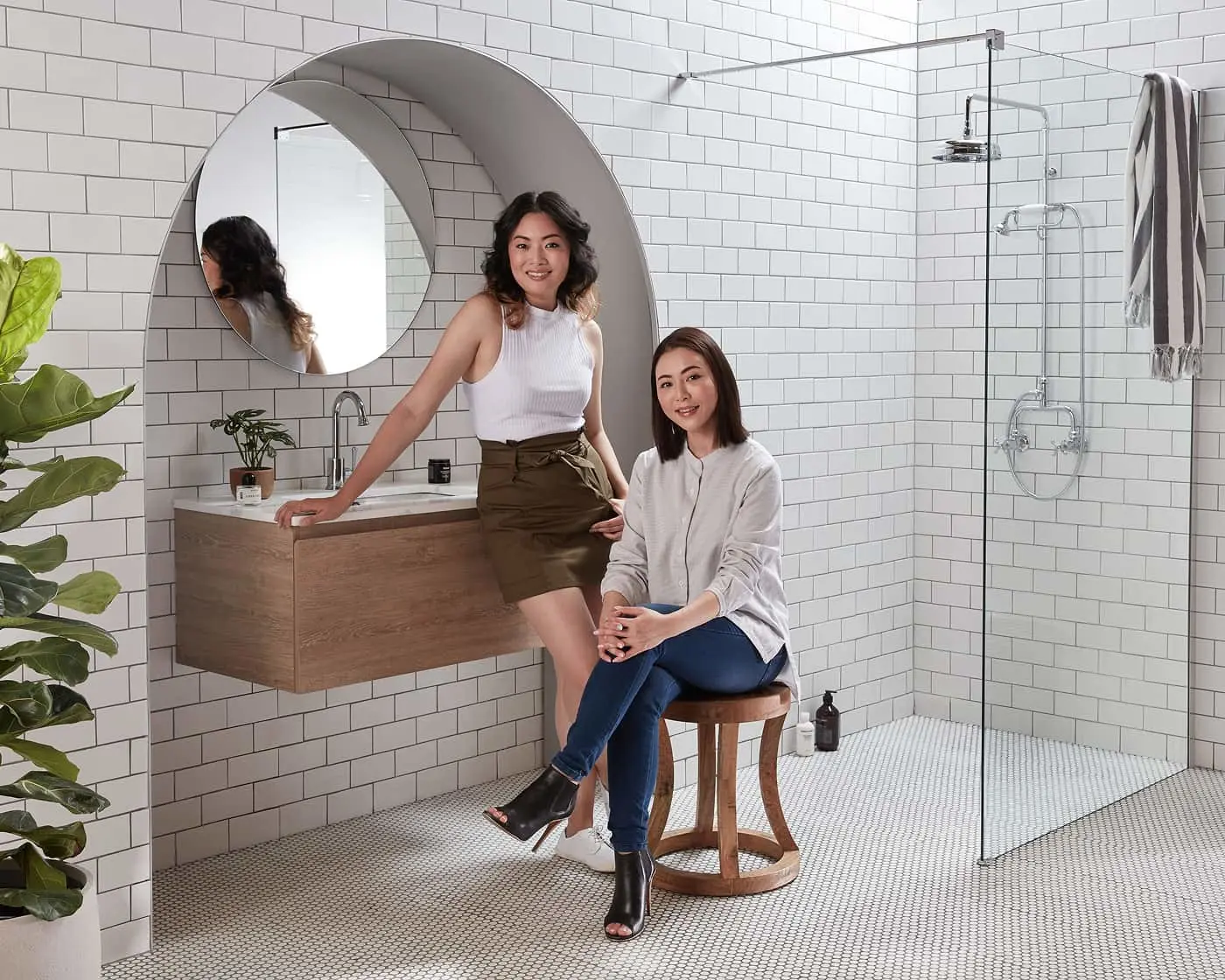 Modern white-tiled bathroom with floating wooden vanity, round mirror, glass shower, and two women posing by the sink.