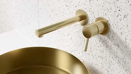 Gold wall-mounted bathroom faucet with matching sink basin against a white speckled wall surface.