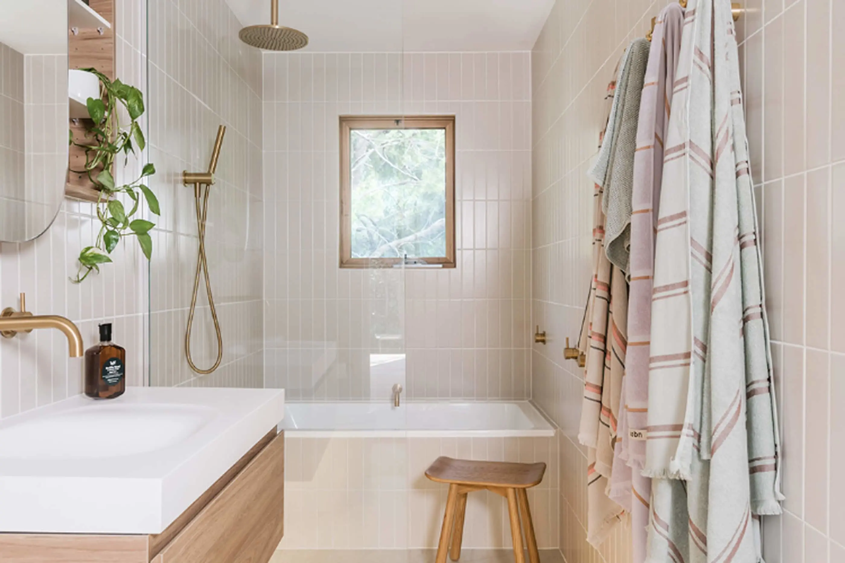 Modern bathroom with cream tiles, brass fixtures, bathtub under window, hanging plant, and pastel towels.