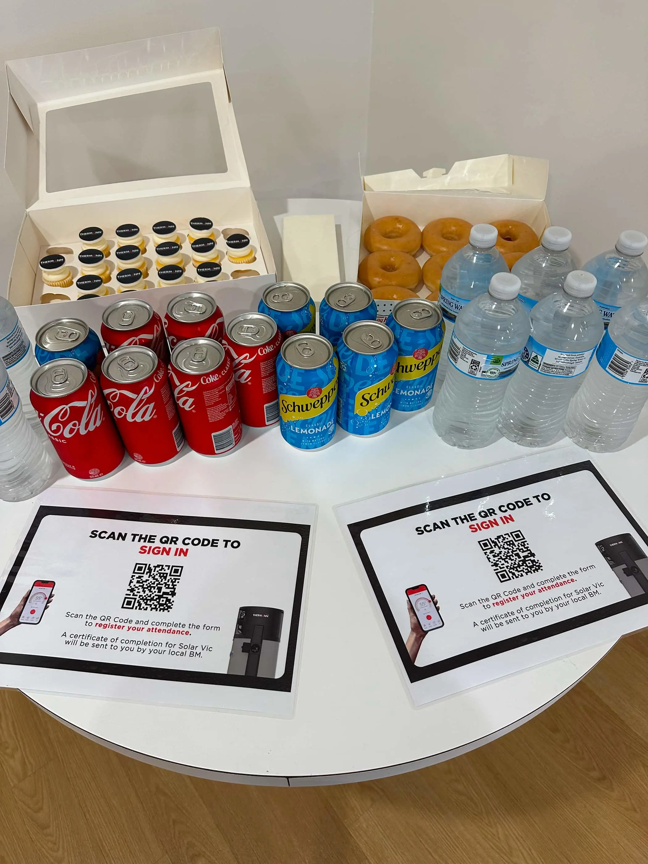 Refreshment table with cupcakes, donuts, Coca-Cola, Schweppes, water bottles, and QR code sign-in sheets.