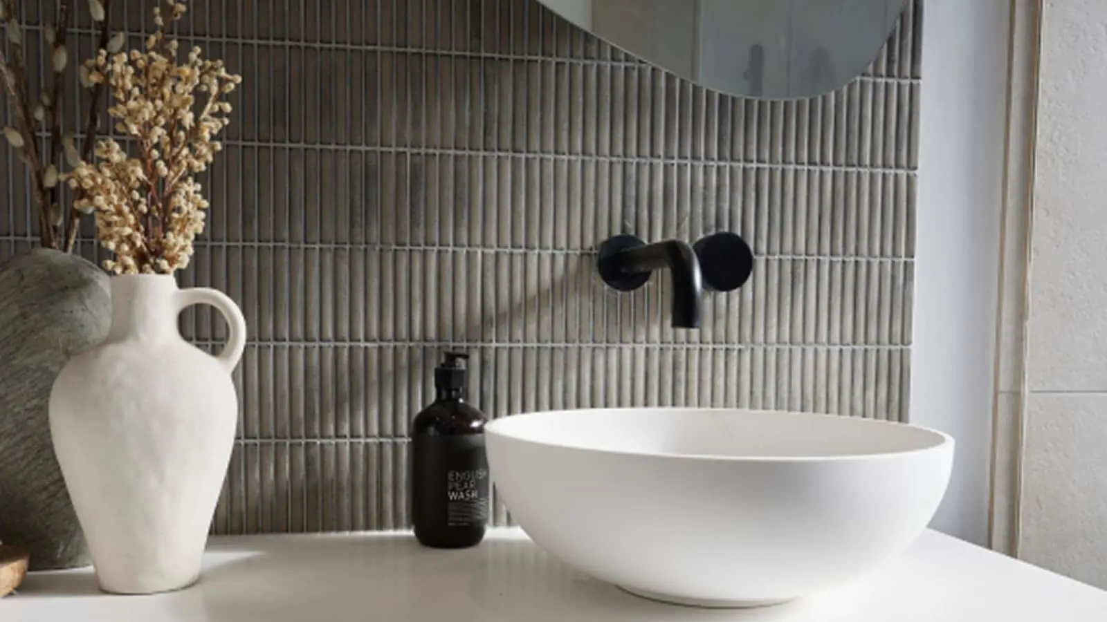 Modern bathroom with white vessel sink, black wall-mounted faucet, gray textured tile backsplash, and decorative white vase.
