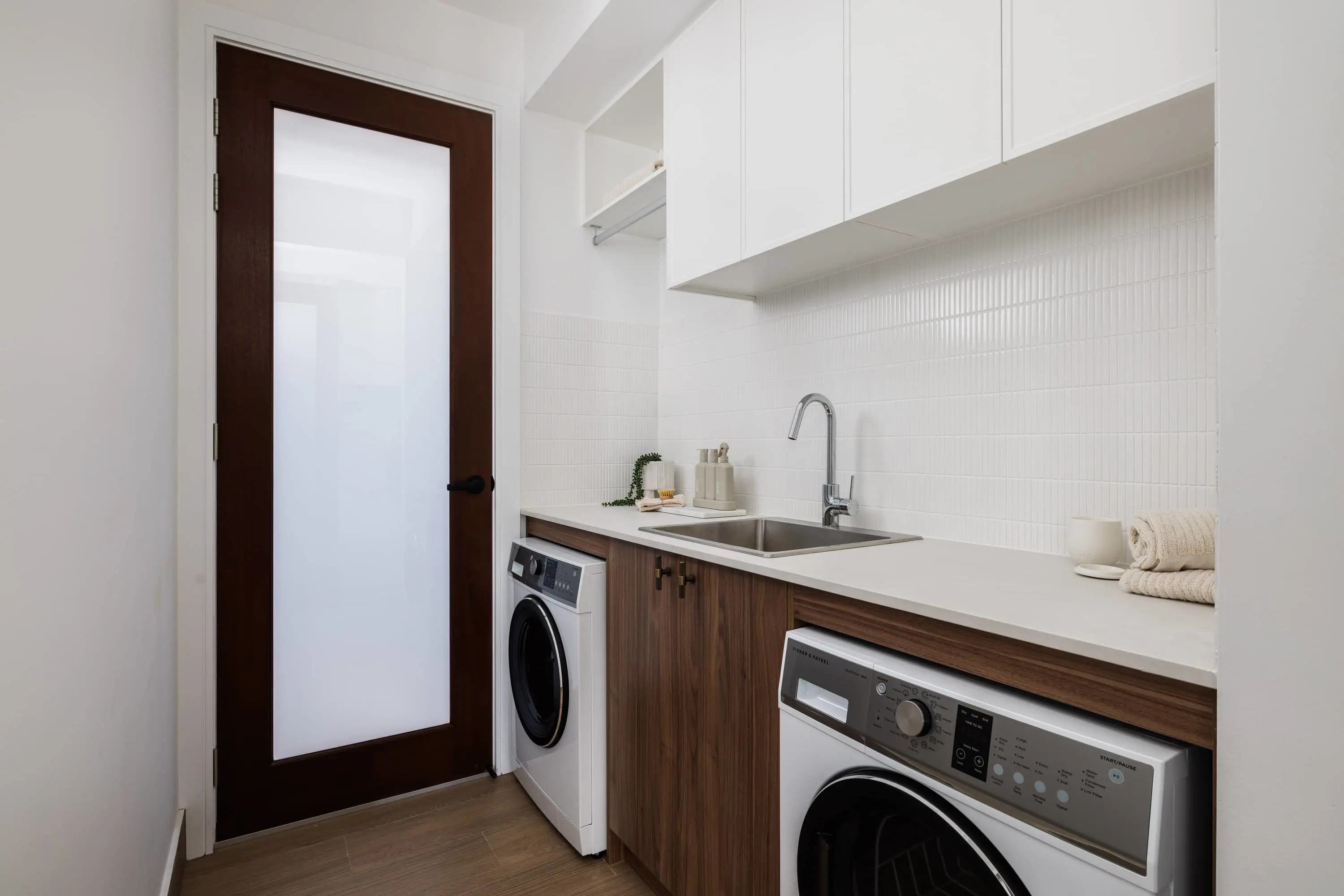 Modern laundry room with white countertop, wooden cabinets, two washing machines, and a frosted glass door.