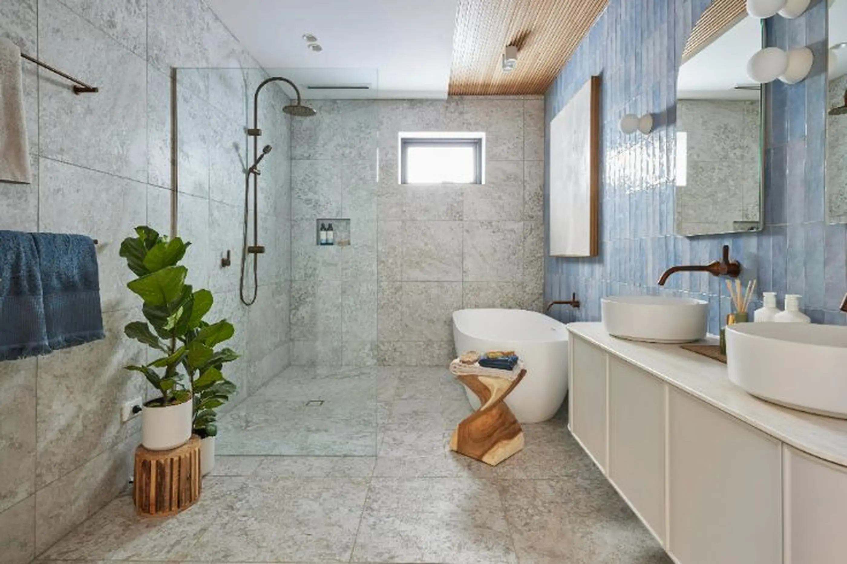 Modern bathroom with marble tiles, walk-in shower, freestanding tub, double vessel sinks, and a green plant on wooden stand.