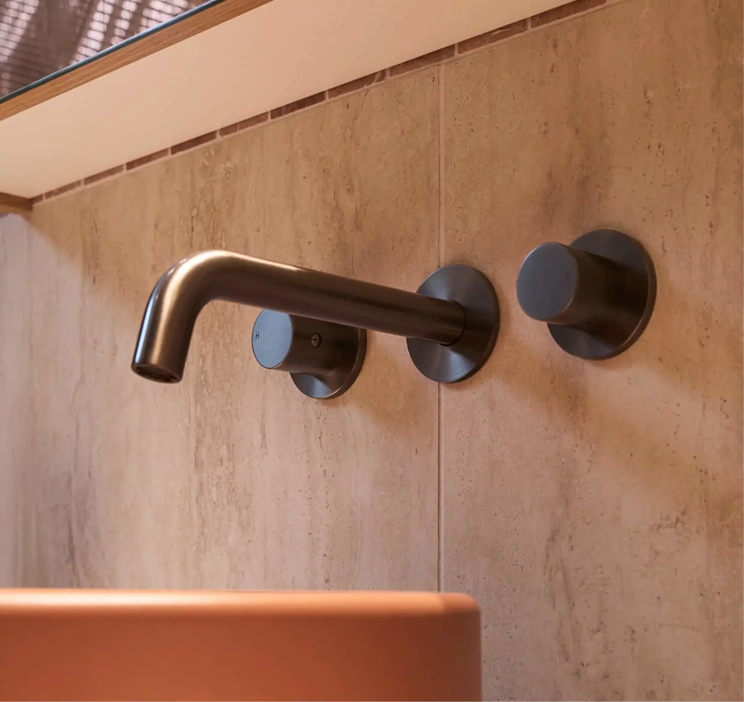 Modern wall-mounted faucet with bronze finish and round handles on beige stone tile wall above terracotta sink.