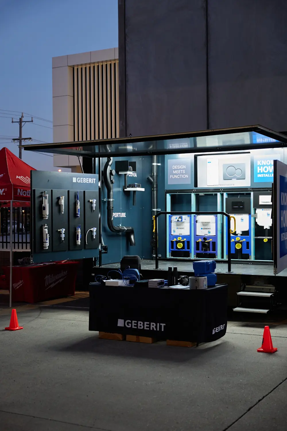 Geberit outdoor product display stand at dusk showing plumbing components, toilet frames, and a branded front desk.