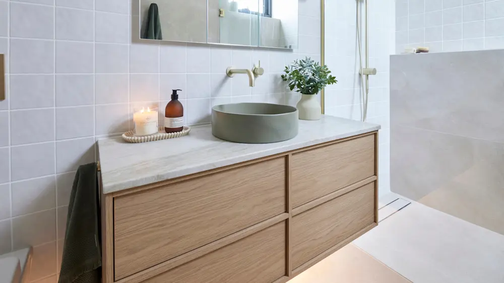 Modern bathroom with wooden vanity, round green sink, brass faucet, lit candle, and small plant in white pot.