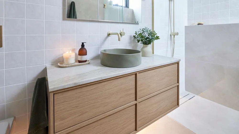 Modern bathroom with wooden vanity, round green sink, brass faucet, lit candle, and small plant in white pot.