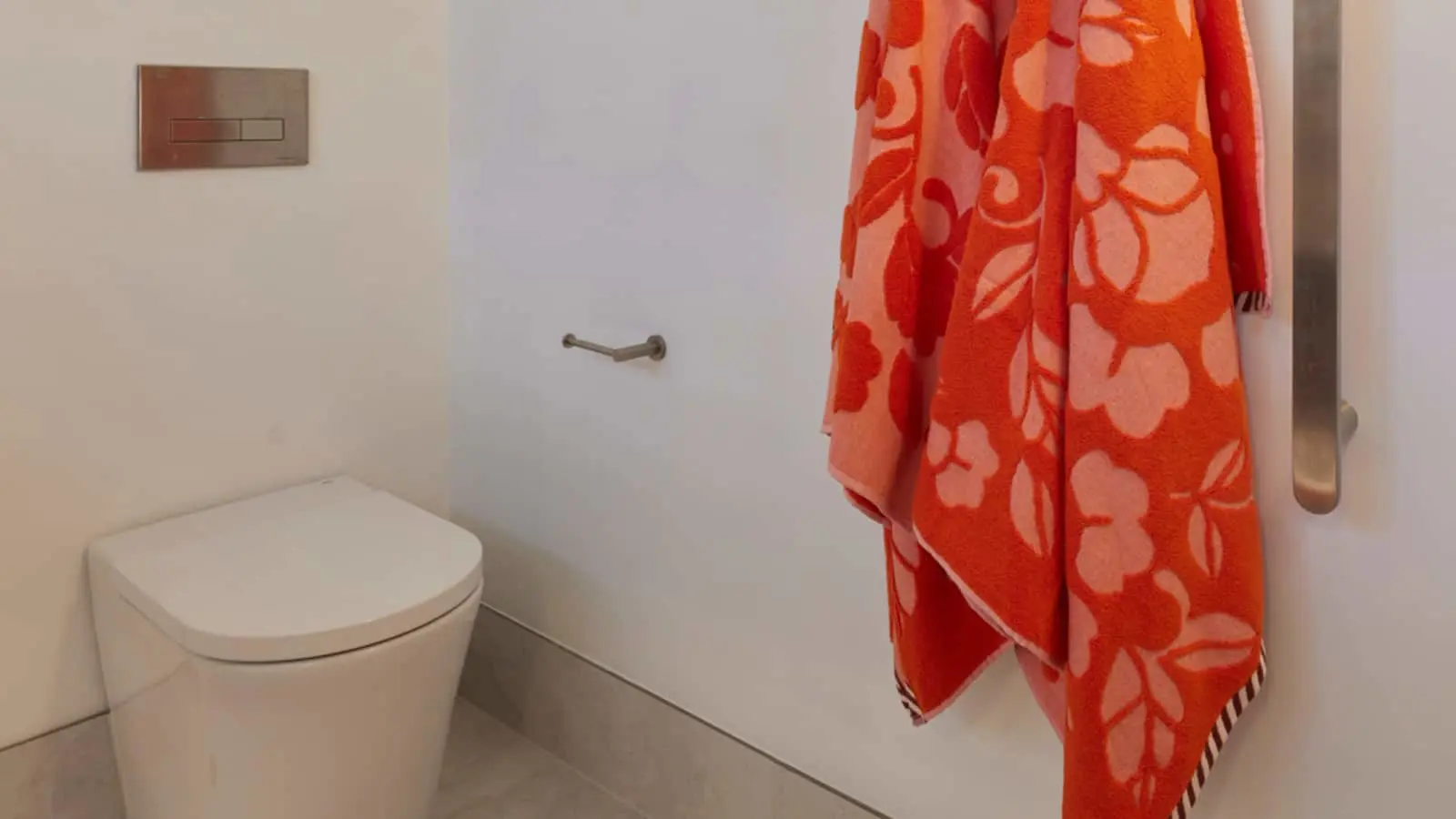 Modern bathroom with white toilet, wall-mounted flush panel, and bright orange floral towel hanging on a metal rail.