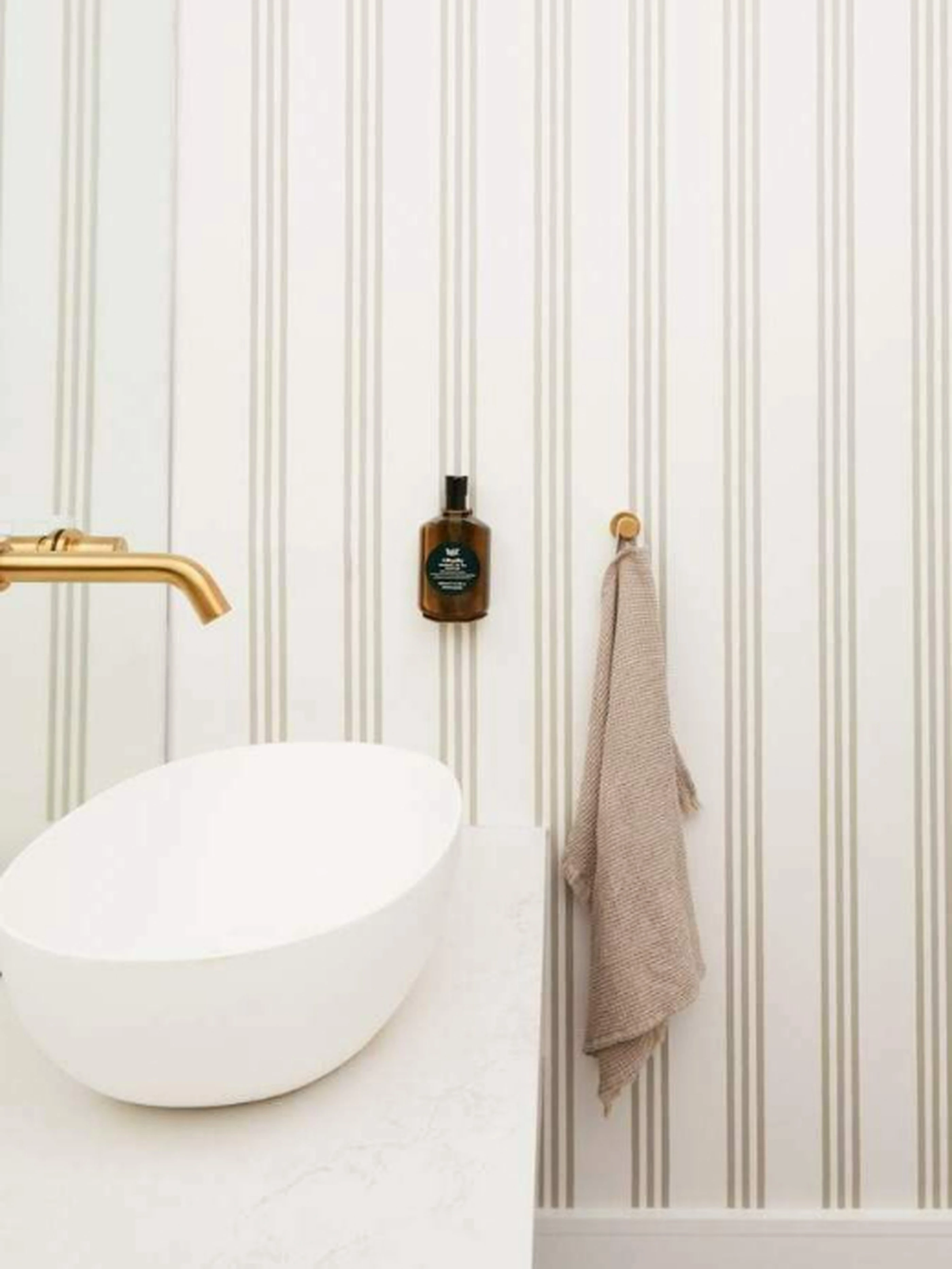 Modern bathroom with white vessel sink, gold faucet, amber bottle, and beige towel against striped wallpaper.