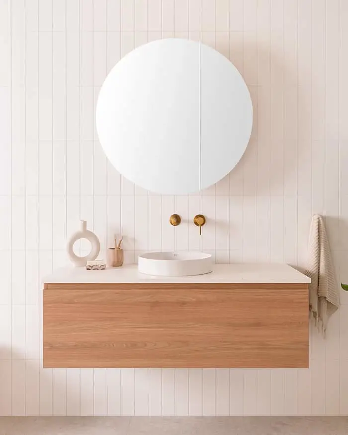 Modern bathroom vanity with wooden cabinet, white sink, round mirror, brass wall faucets, and minimal decor on light walls.