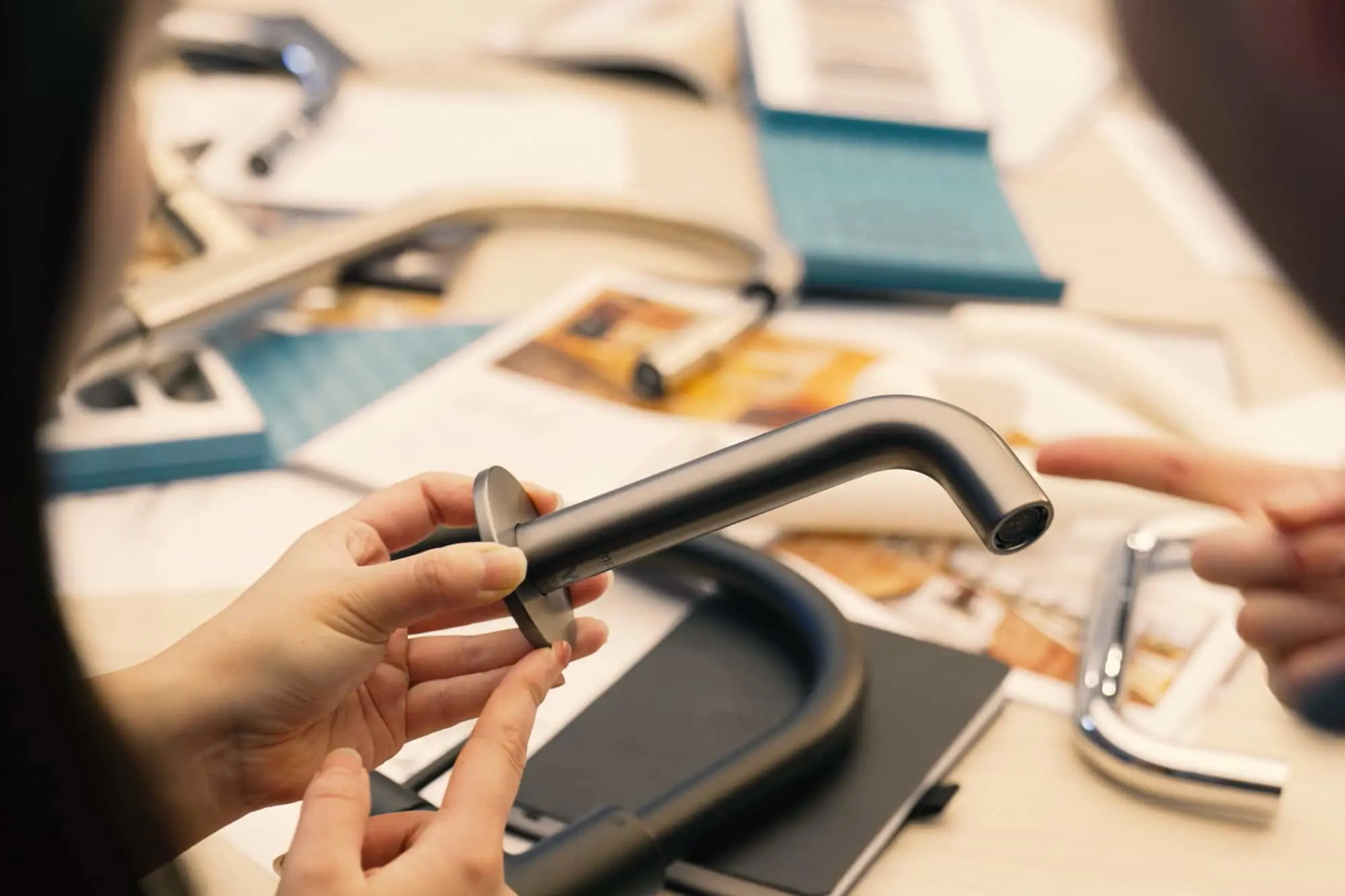 Hands holding a modern silver faucet with design materials and product samples spread on a table in the background.