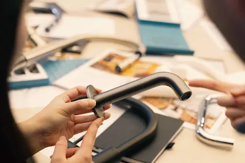 Hands holding a modern silver faucet with design materials and product samples spread on a table in the background.