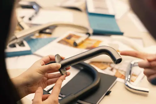 Hands holding a modern silver faucet with design materials and product samples spread on a table in the background.