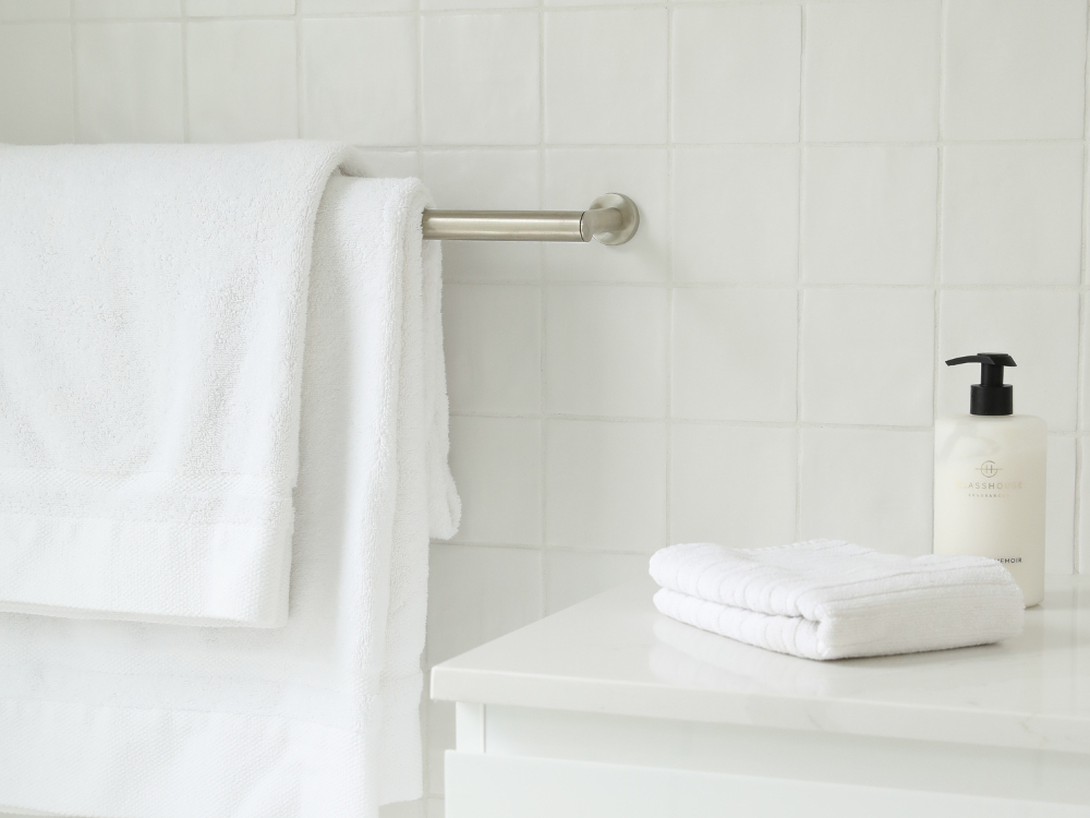 White towels hanging on a metal rack, with folded washcloths and a pump bottle on a counter in a tiled bathroom.