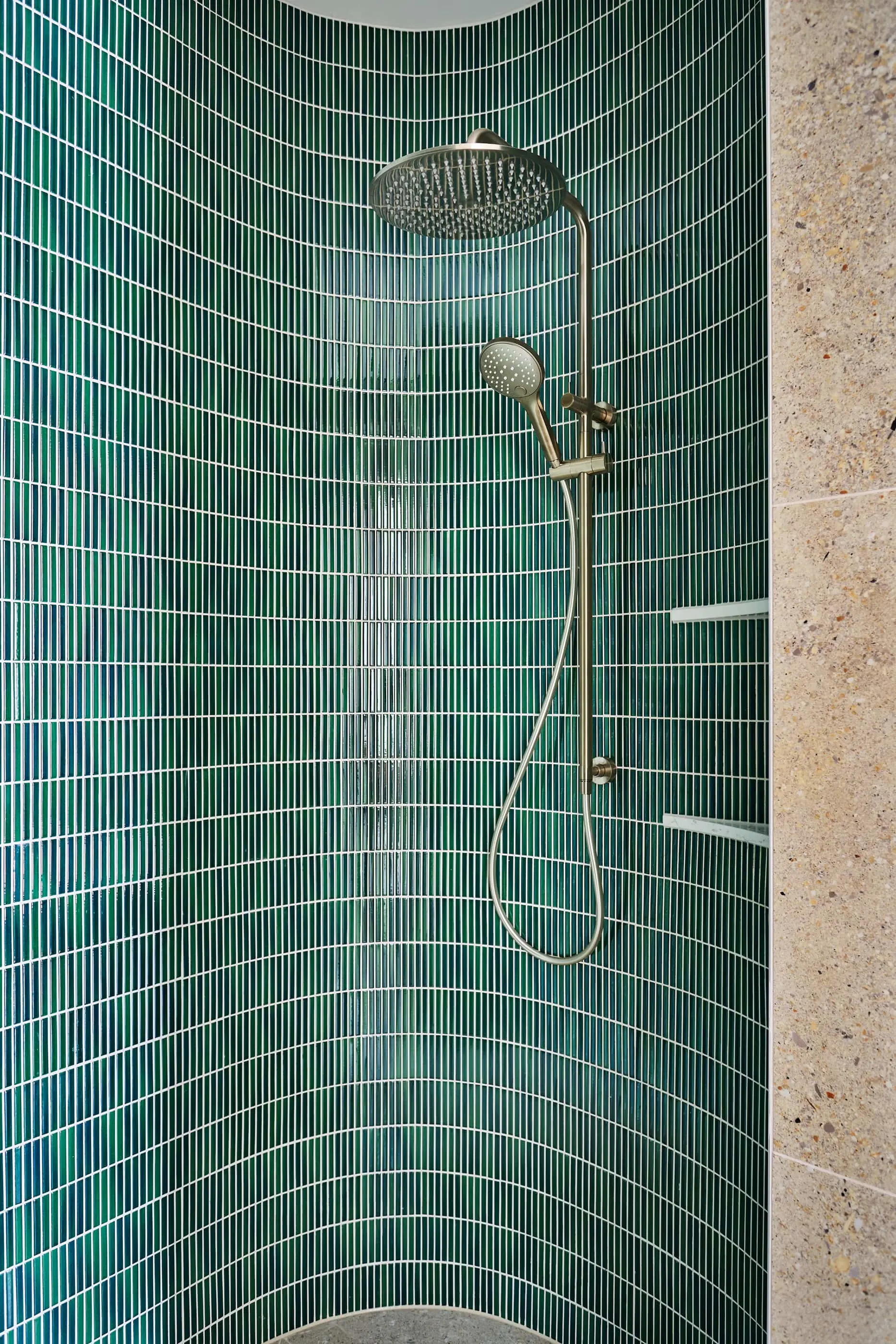 Modern shower with brass fixtures featuring a rainfall showerhead and handheld sprayer against green tiled curved walls.