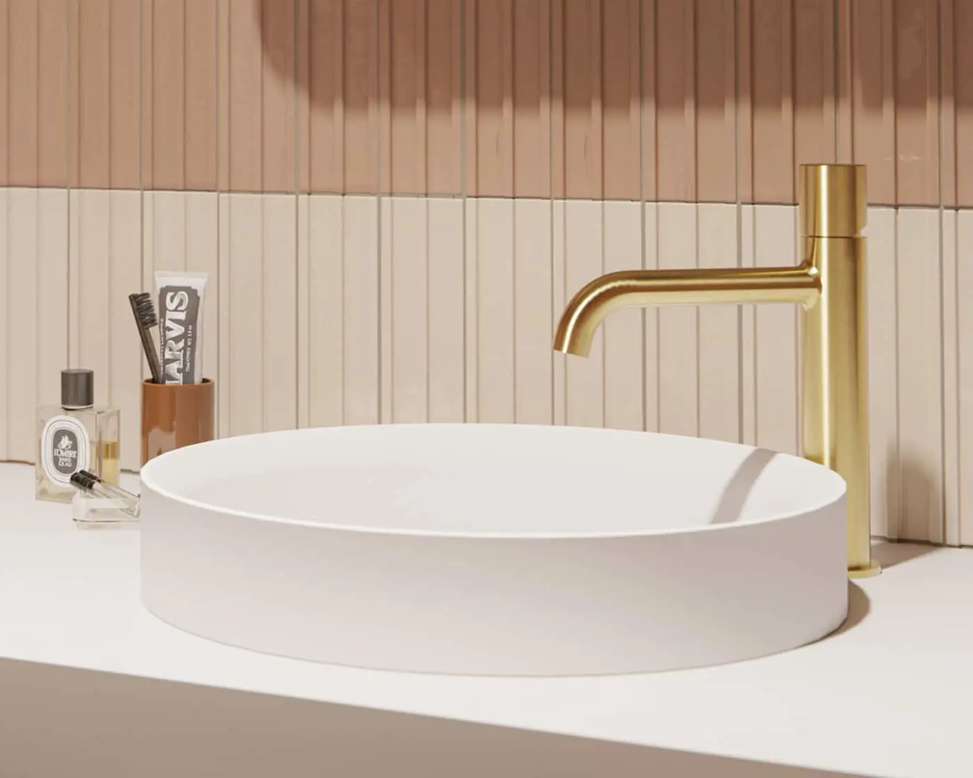 Modern bathroom sink with gold faucet, toothbrush holder, and perfume bottles against pink and cream tiled wall.