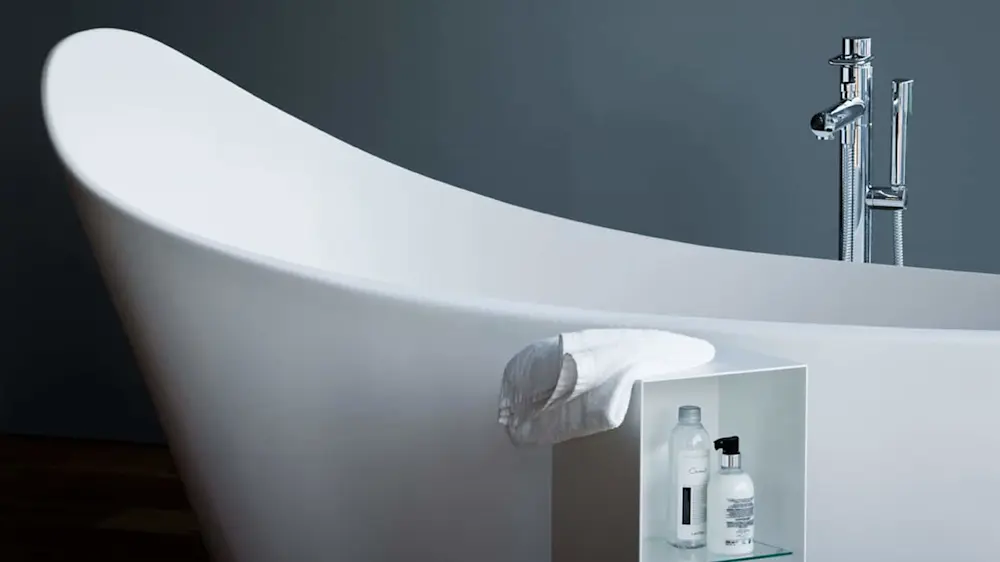 Modern white freestanding bathtub with chrome faucet, small shelf holding bath products, and folded towel.
