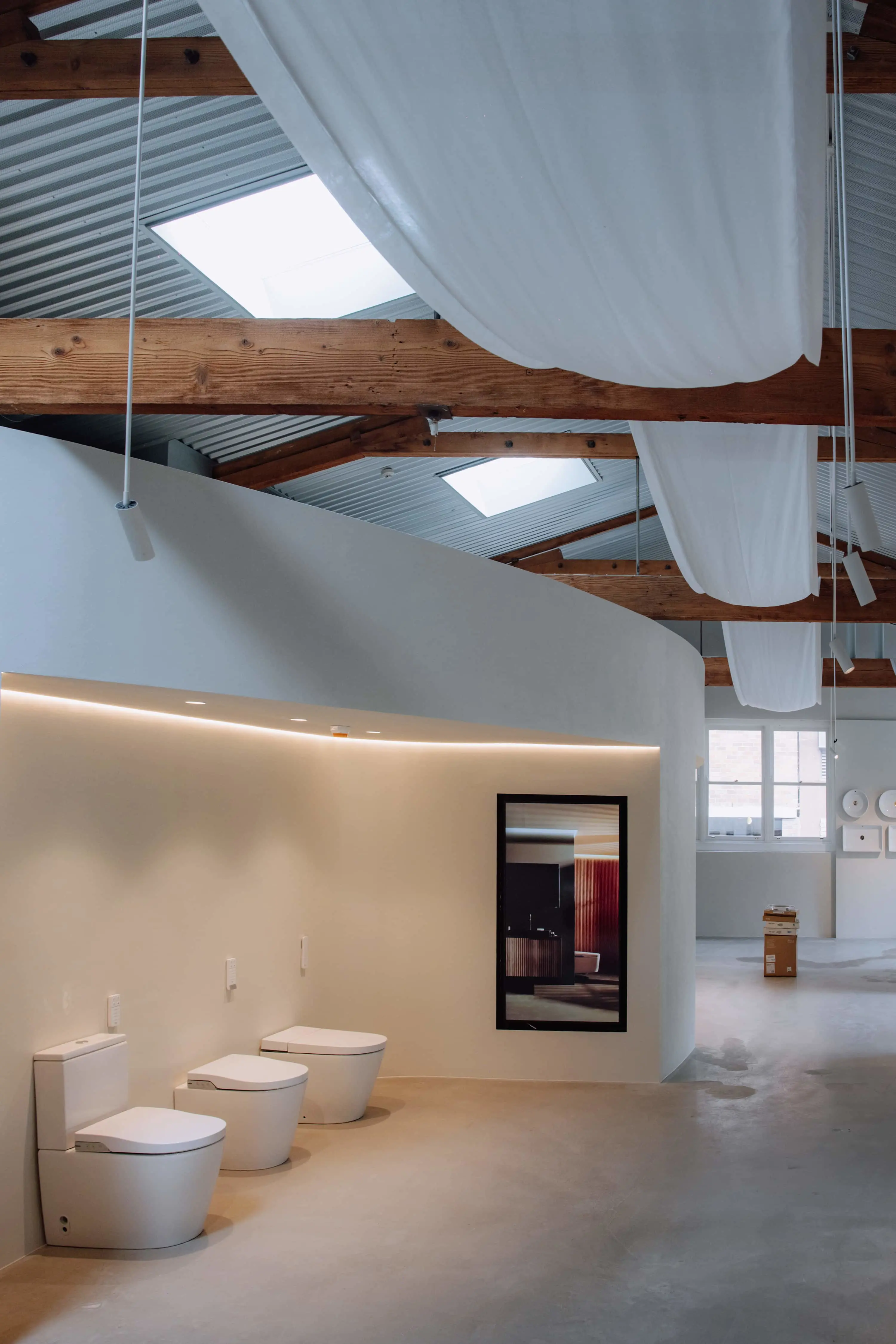Modern bathroom showroom with white toilets displayed against a curved wall, wooden beams overhead, and skylights.