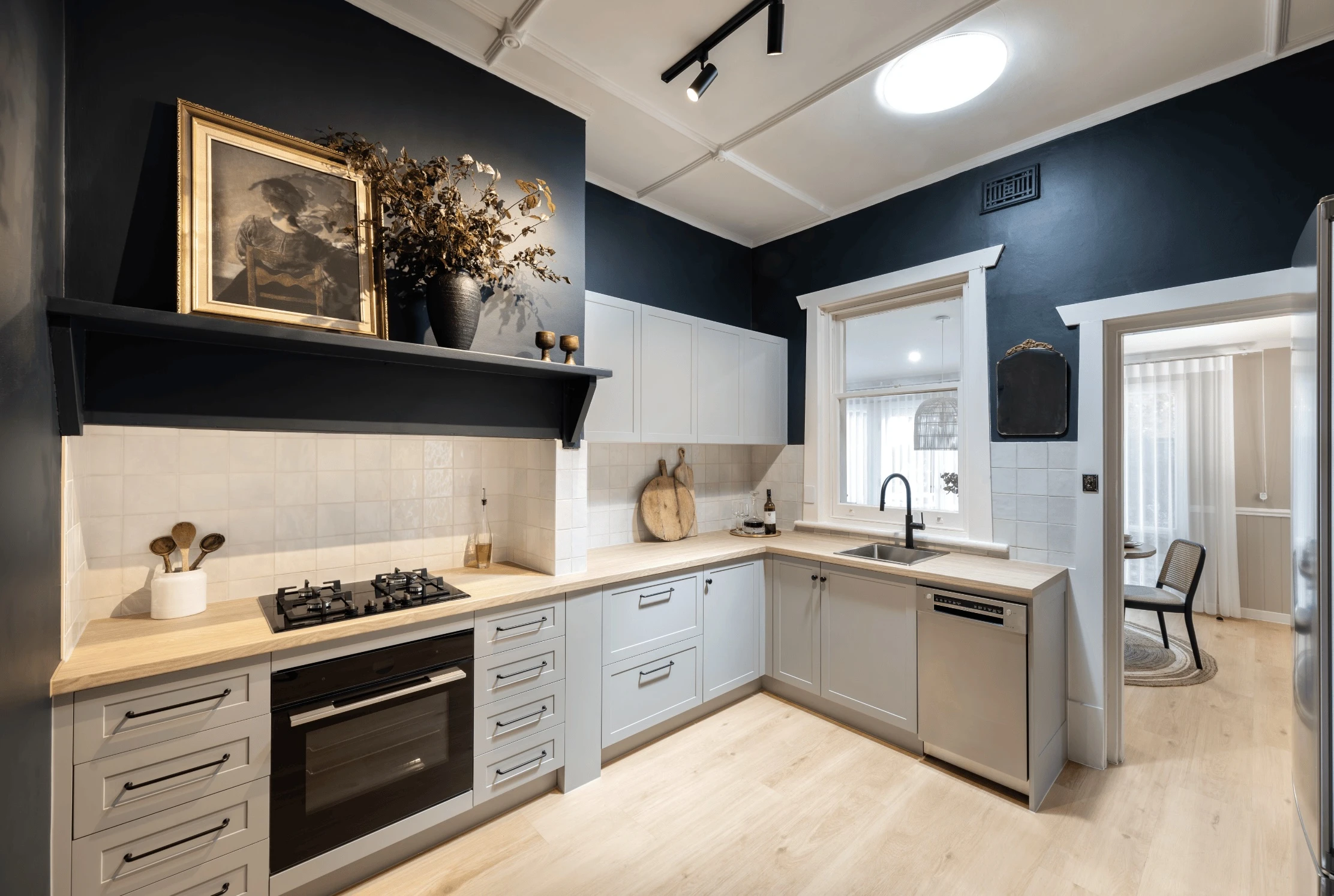Modern kitchen with navy blue walls, white cabinets, wooden countertops, and decorative shelf displaying artwork and dried flowers.