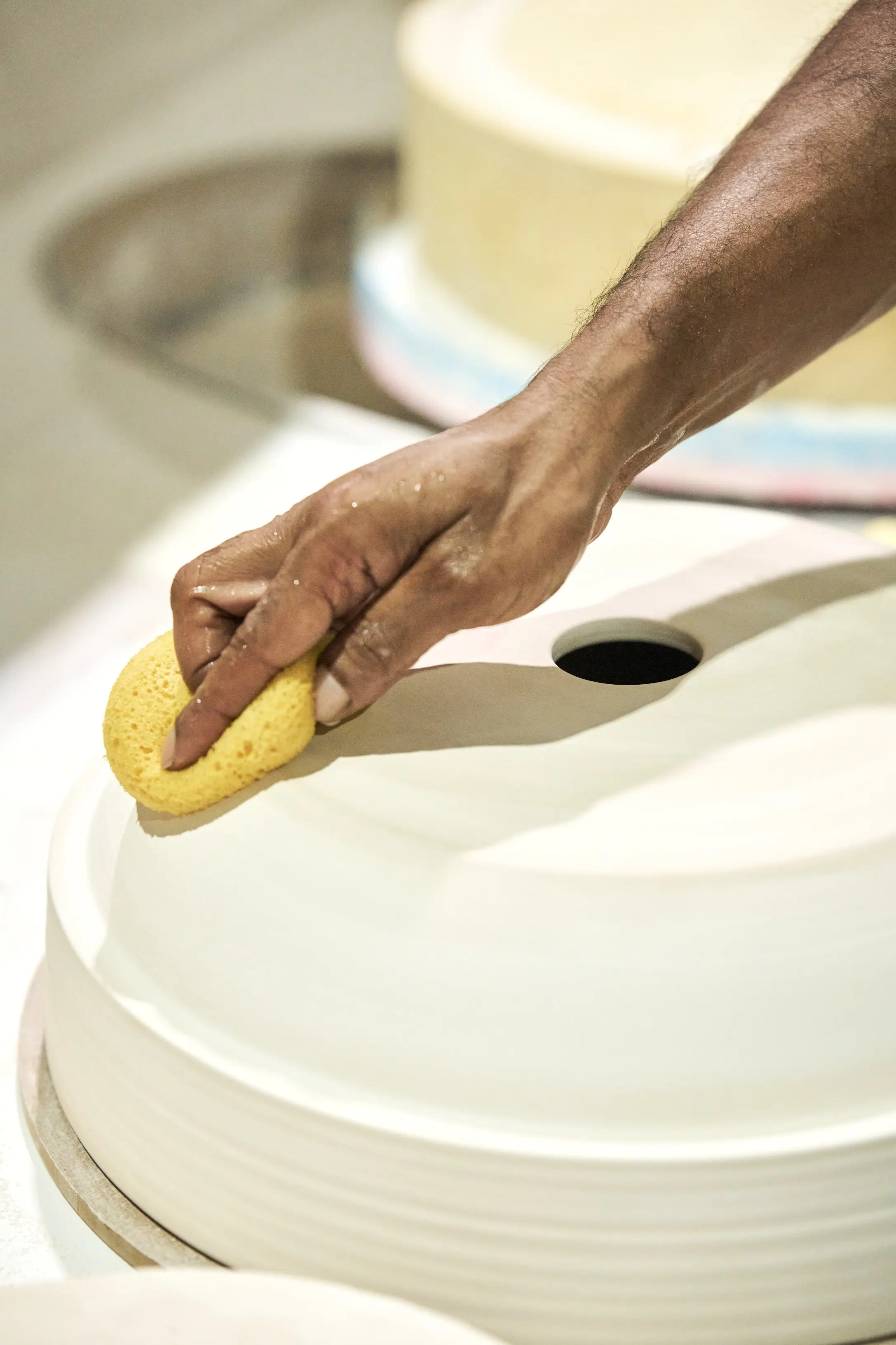 A hand using a yellow sponge to smooth wet clay on a pottery wheel, with a water bowl visible in the background.