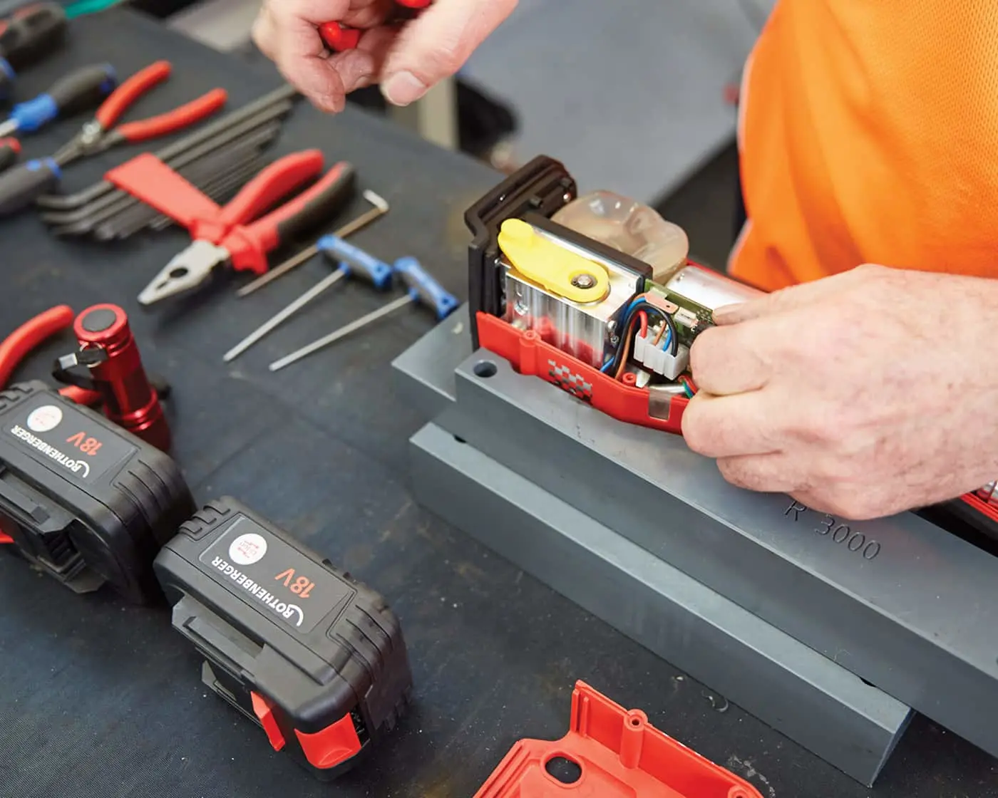 Person in orange shirt repairing an electronic device with tools and battery packs visible on workbench.