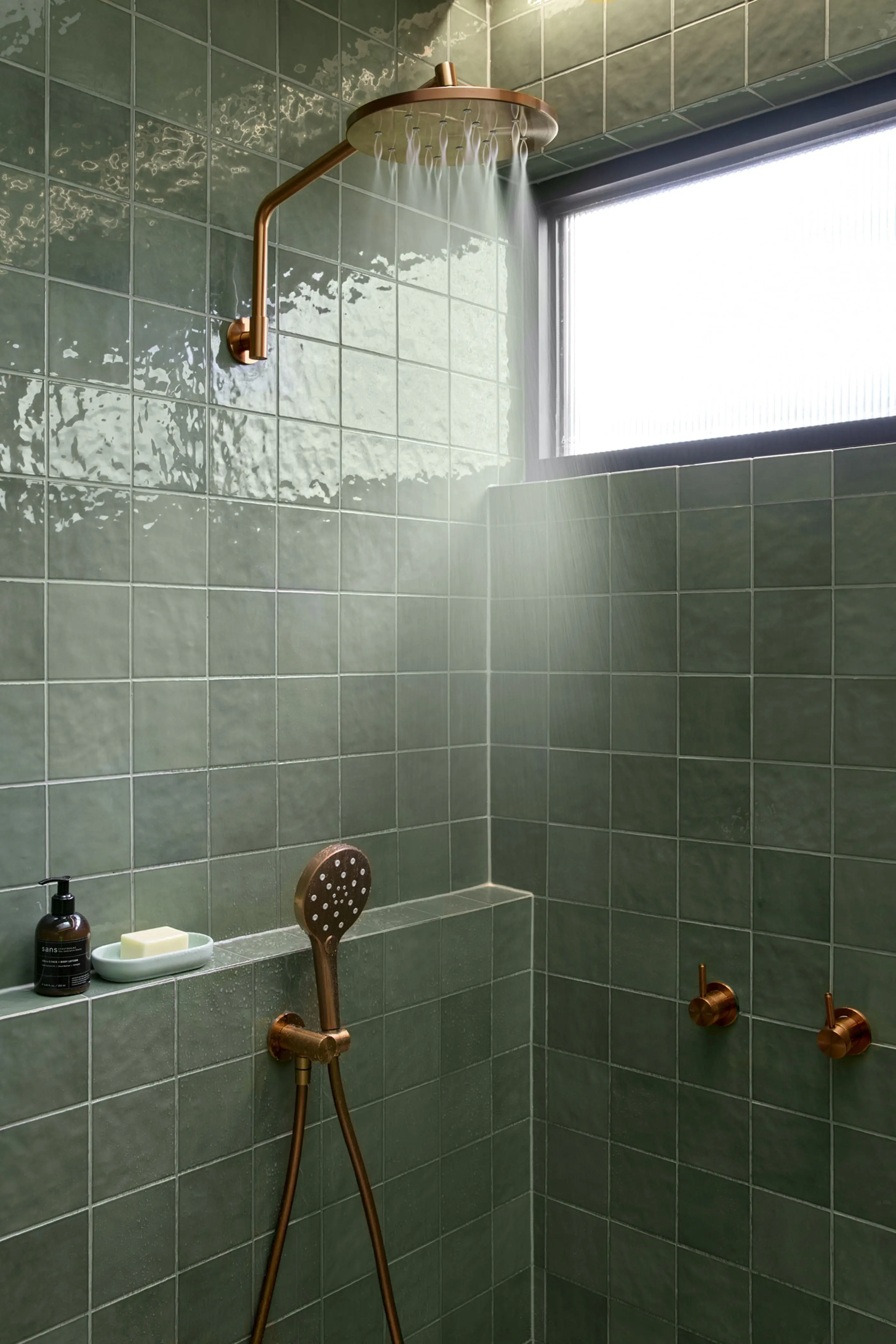 Modern bathroom with green tile walls, copper rainfall showerhead and handheld shower, small window, and built-in shelf.