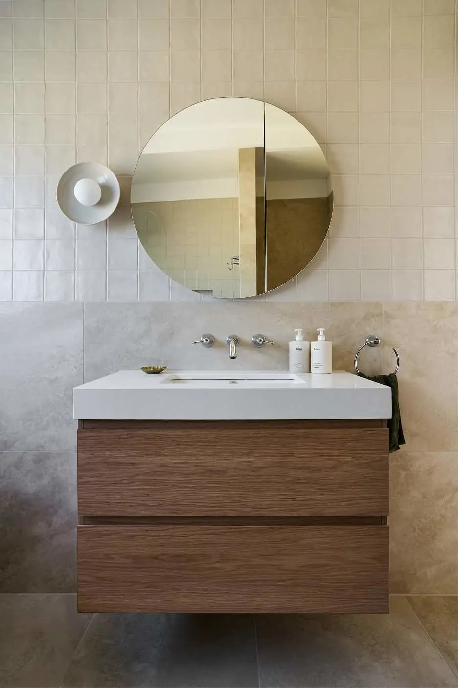 Modern bathroom with wooden vanity, white sink, round mirror, and beige tiled walls. Two product dispensers sit on the countertop.