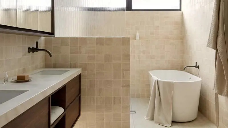 Neutral-toned bathroom with beige square tiles, a freestanding white bathtub, dark wood vanity, and matte black wall-mounted faucets.