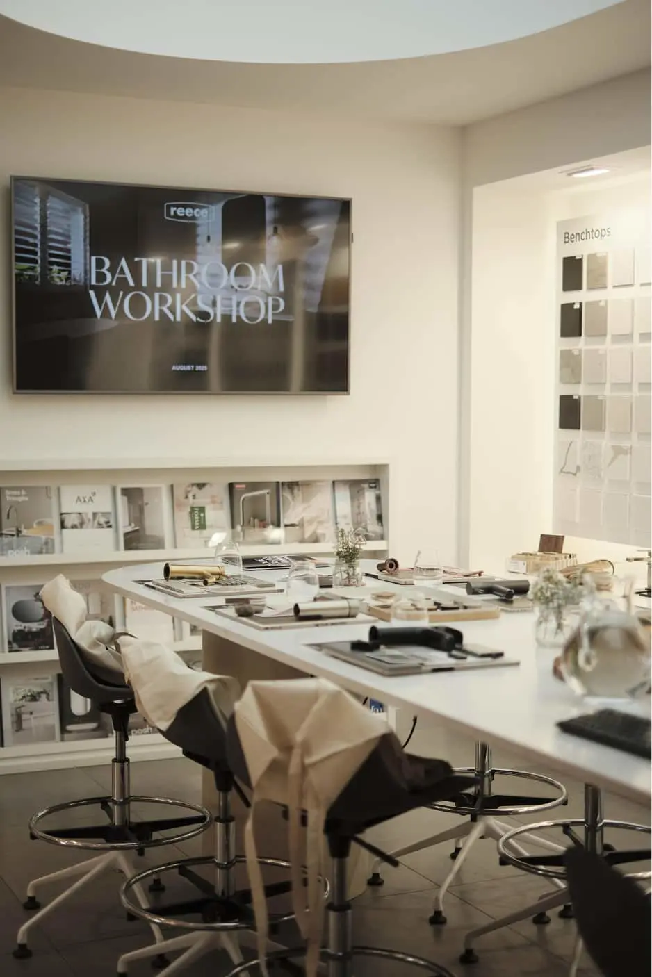 Bathroom workshop room with white table, high chairs, design materials, wall-mounted screen, and benchtop samples display.