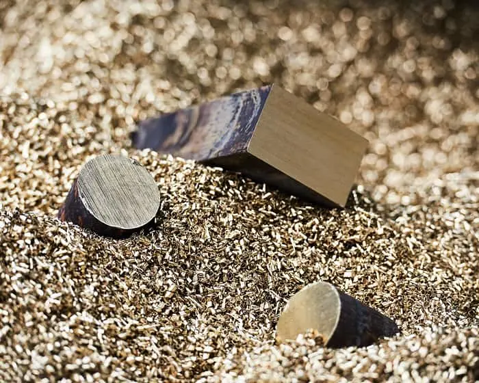 Gold metal pieces including a cube and circular discs resting on shimmering gold particles.
