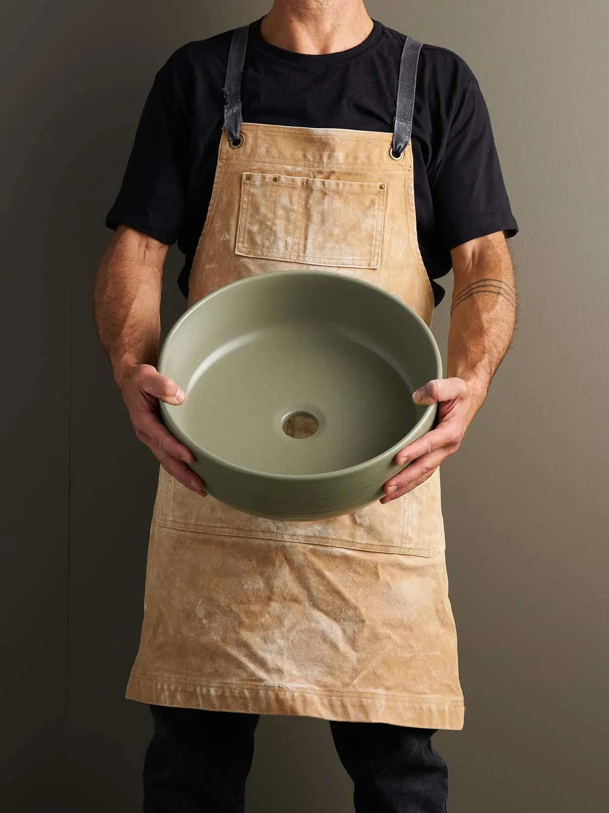 Person in tan apron and black shirt holding a round sage green ceramic basin or sink.