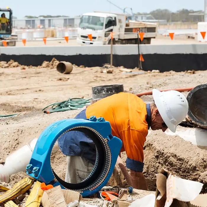 Construction worker in orange safety gear installing plumbing at a building site with trucks and safety markers visible.