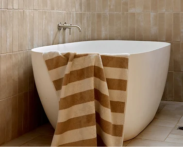 Modern white freestanding bathtub with brown striped towel draped over edge against beige tiled wall