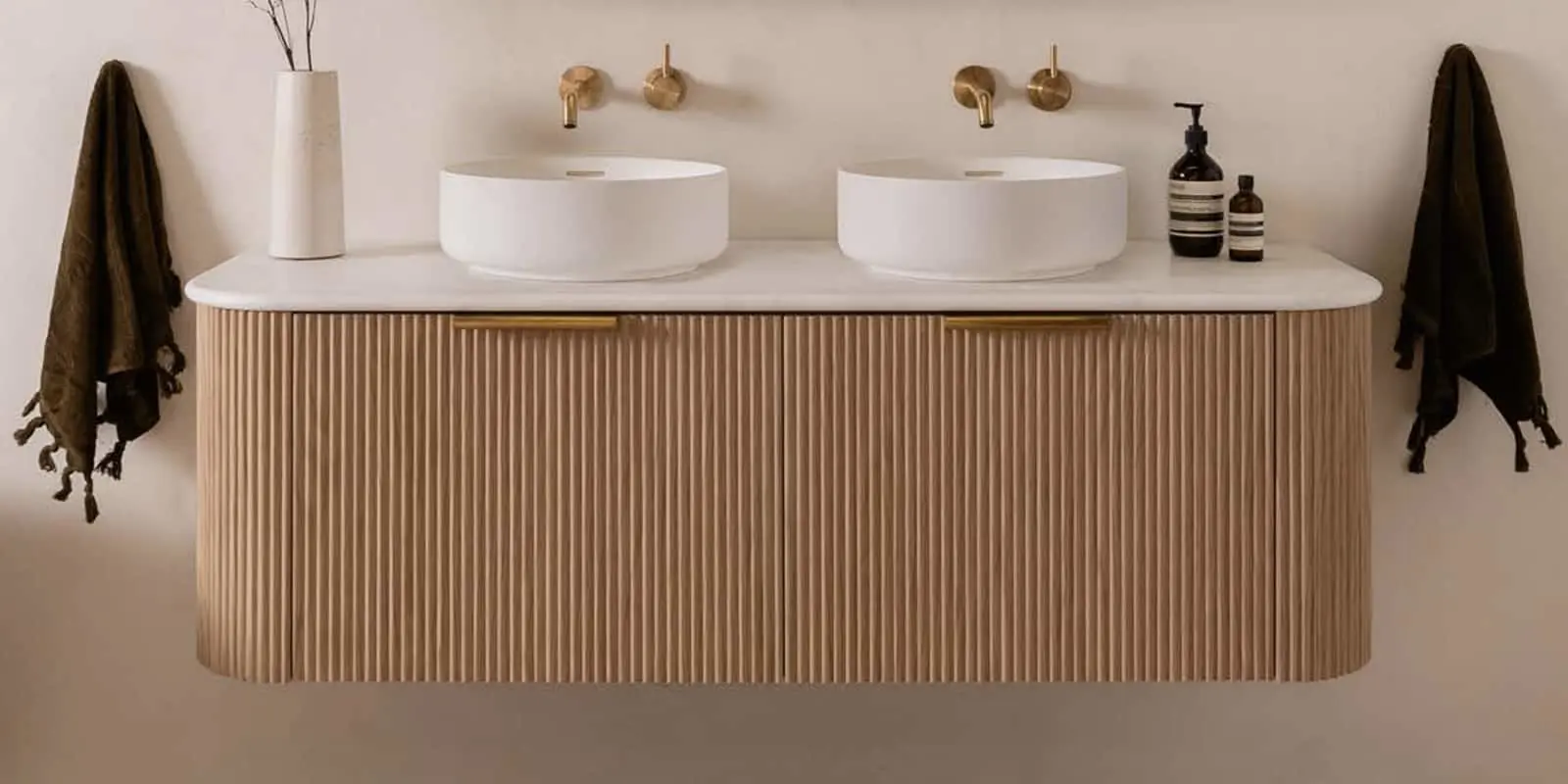 Modern bathroom vanity with wooden ribbed cabinet, dual white vessel sinks, brass faucets, and dark towels on either side.