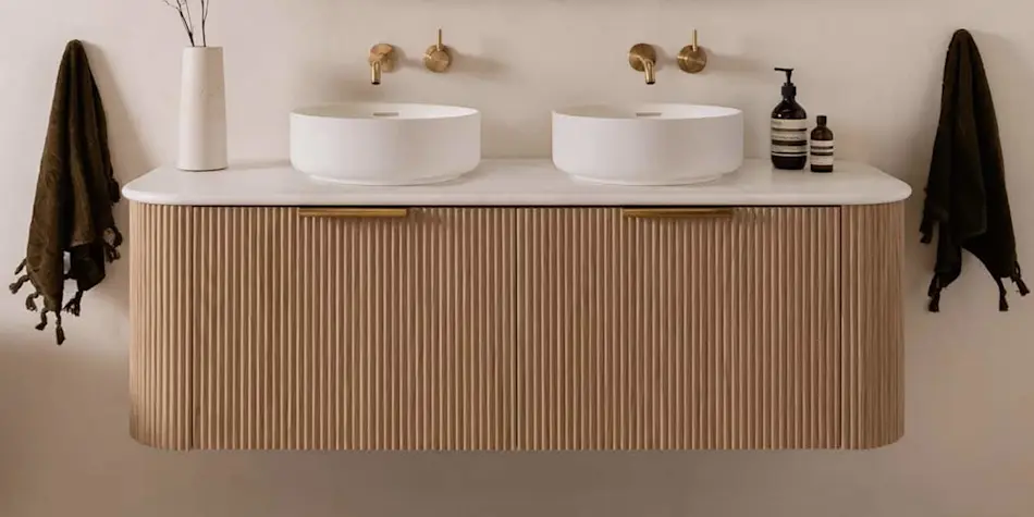 Modern bathroom vanity with wooden ribbed cabinet, dual white vessel sinks, brass faucets, and dark towels on either side.