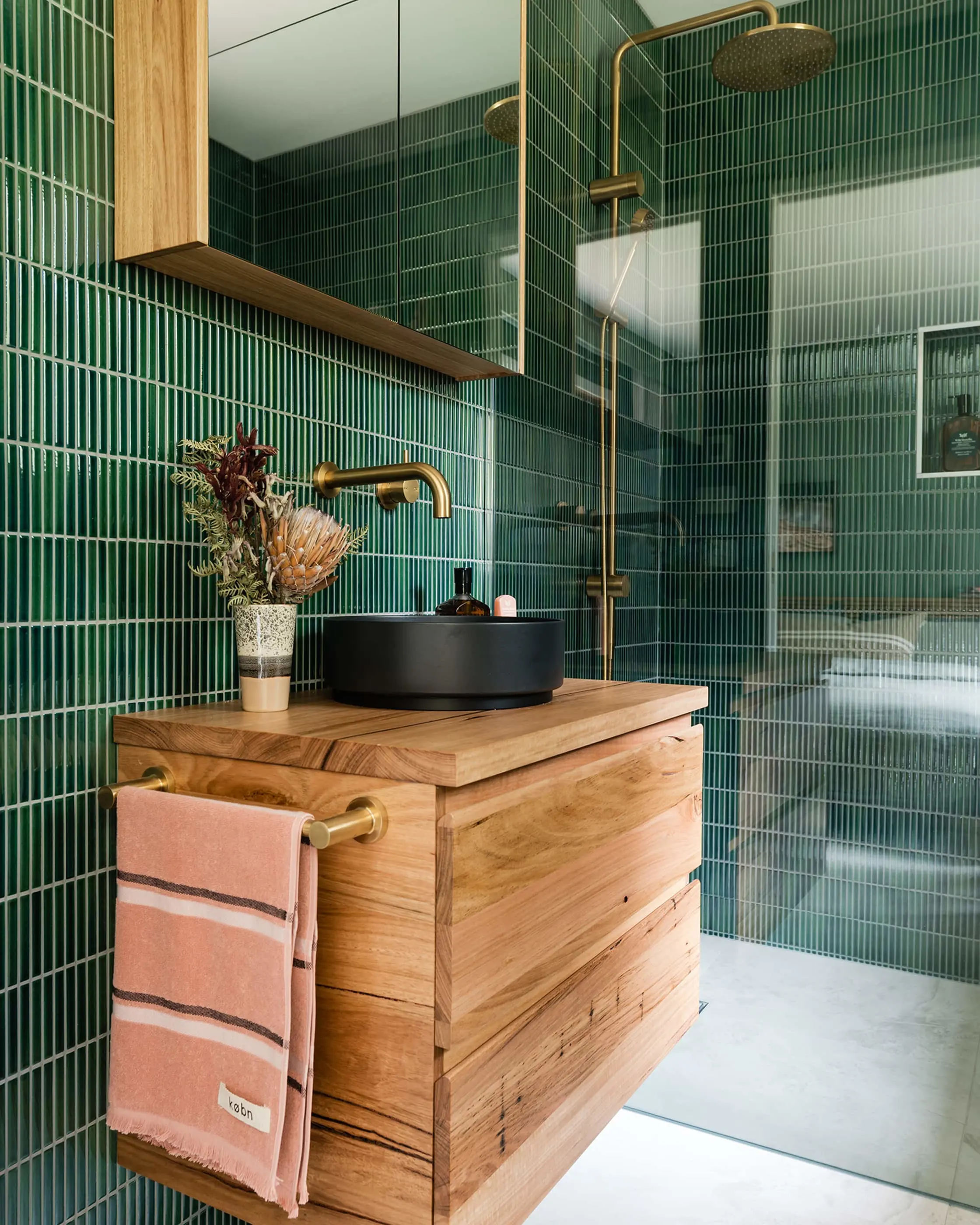 Modern bathroom with green tile walls, wooden vanity with black basin, brass fixtures, and pink towel hanging on side.