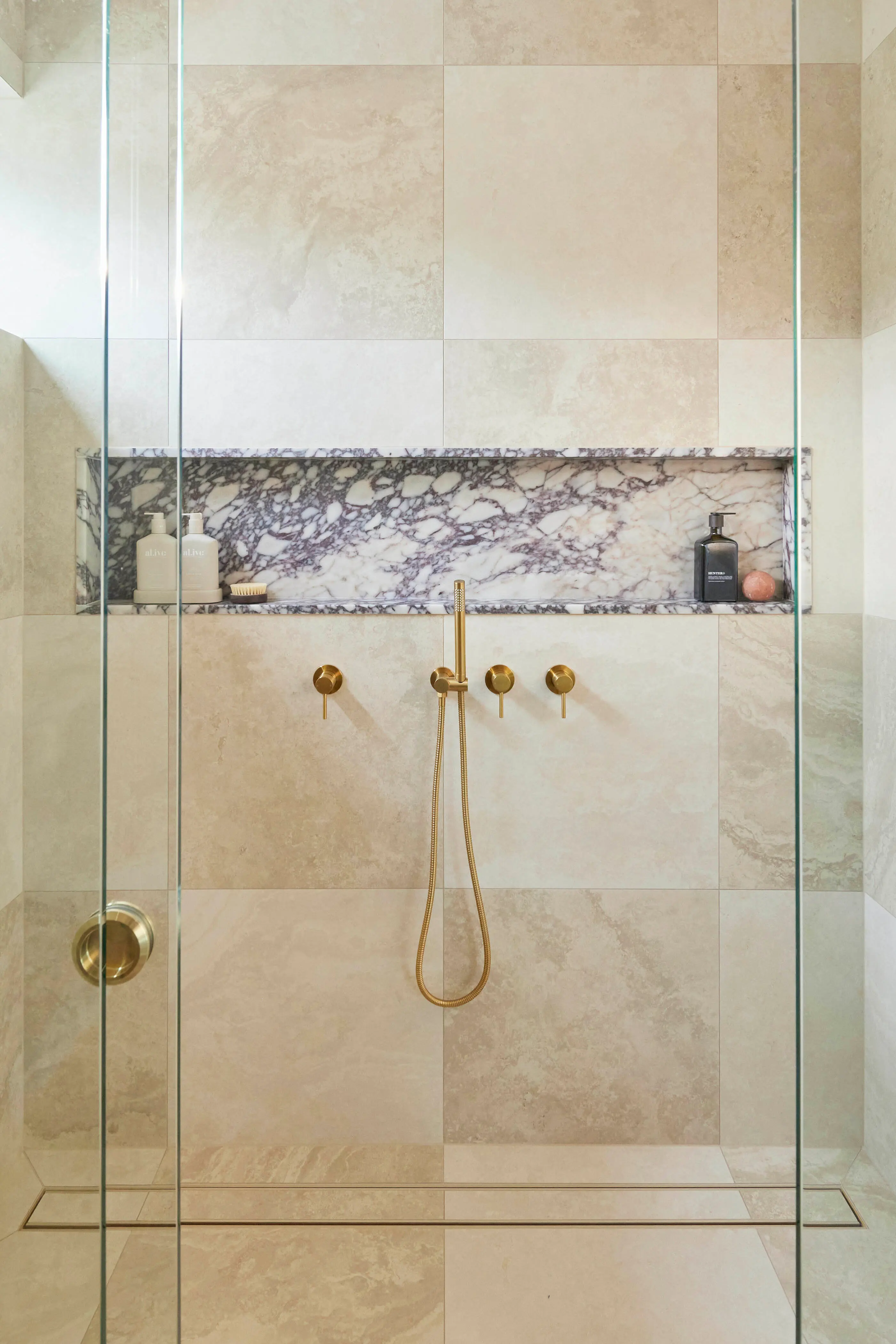 Modern glass shower with beige tile walls, marble recessed shelf, and brass fixtures including handheld showerhead.