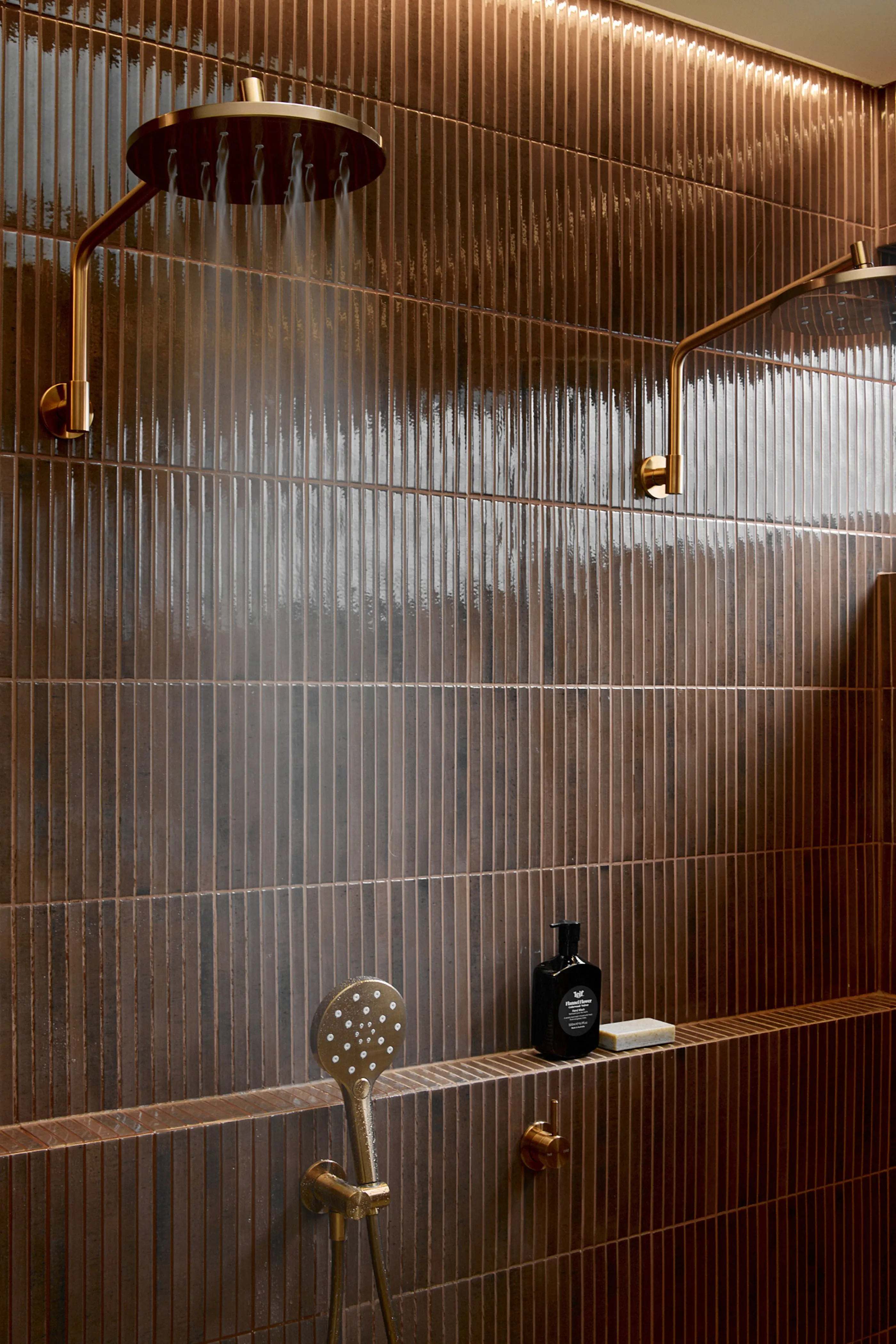 Modern shower with bronze fixtures, vertical tiled walls, rainfall showerhead, handheld sprayer, and black toiletry bottle on shelf.