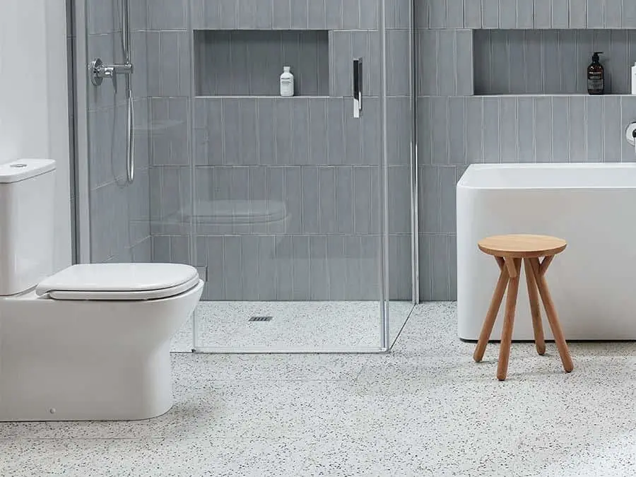 Modern bathroom with gray tile walls, white toilet, glass shower enclosure, bathtub, and wooden stool on terrazzo floor.