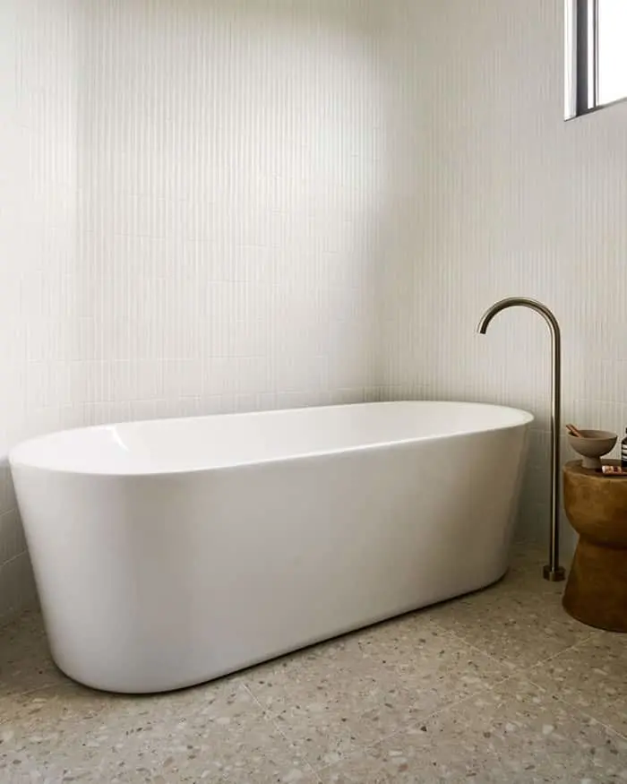 Modern white freestanding bathtub with brass faucet in minimalist bathroom with textured walls and stone floor.