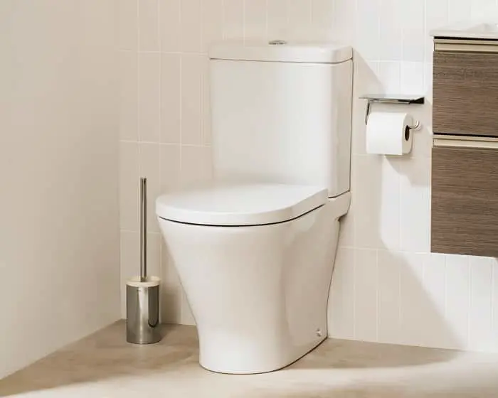 White toilet in a bathroom with white tiled walls, toilet brush holder, and wooden cabinet drawers.