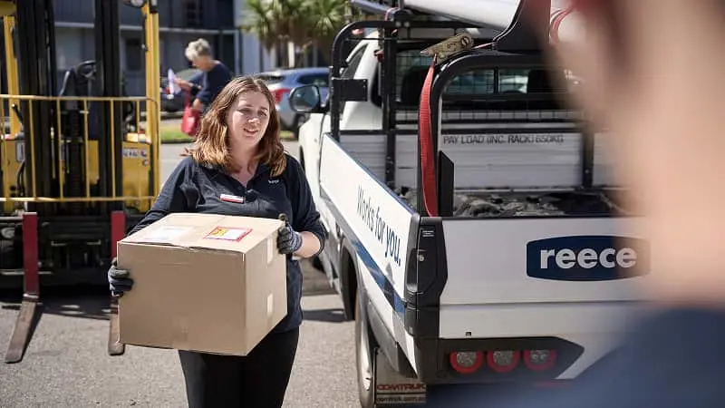 Delivery worker in navy uniform carrying a cardboard box next to a delivery truck with "Works for you" signage.