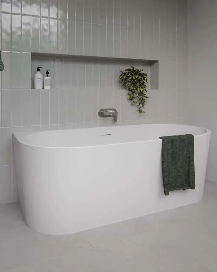 White freestanding bathtub against grey tiled wall with wall-mounted tap, niche shelf with bottles and green plant, dark towel draped over tub.