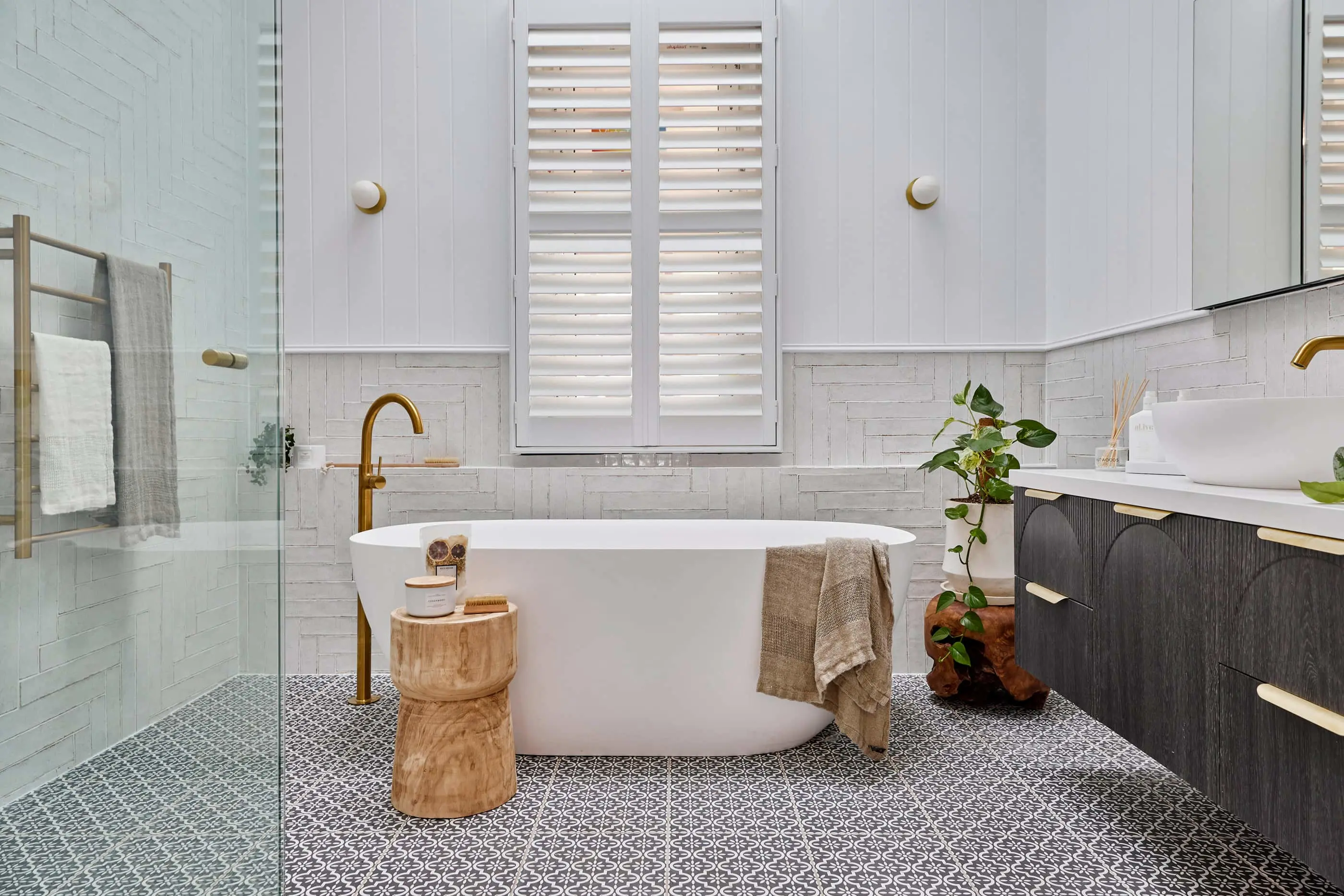 Modern white bathroom with freestanding tub, wooden stool, patterned floor tiles, and white plantation shutters.