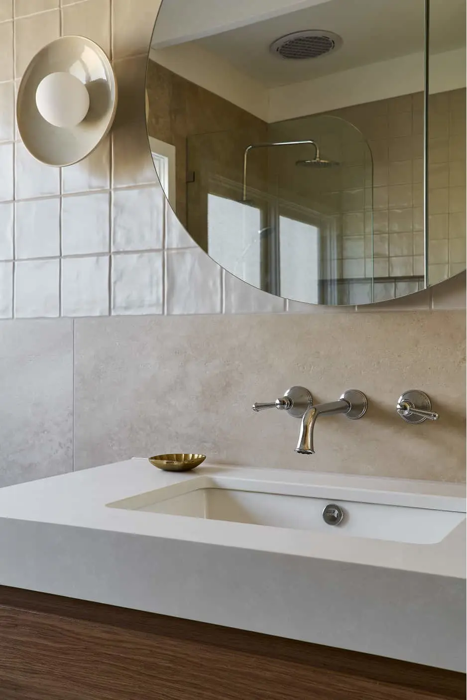 Modern bathroom with white sink, wall-mounted chrome faucet, round mirror, and circular wall sconce on beige tiled walls.