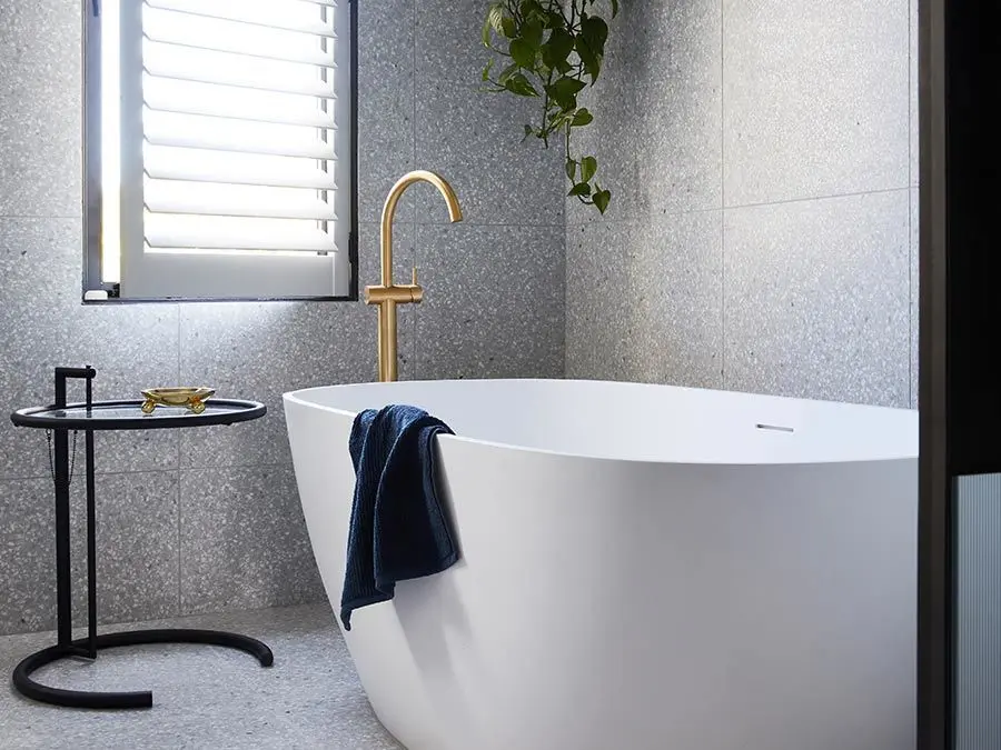 Modern bathroom with white freestanding tub, gold faucet, gray tile walls, window with blinds, and hanging plant.